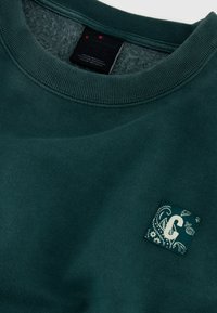 Zielona bluza z okrągłym dekoltem, wykonana z teksturowanej tkaniny, z małą prostokątną naszywką z wzorem paisley.