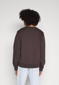 Dunkelbrauner Pullover mit langen Ärmeln, Rundhalsausschnitt und gerippten Bündchen. Weicher Stoff, minimalistisches Design, getragen mit hellblauen Jeans.