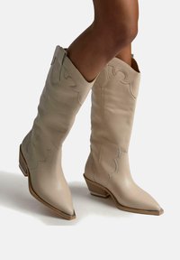Botas de cuero beige hasta la rodilla con puntera puntiaguda, diseño ondulado y un pequeño tacón angular. Textura suave con un sutil detalle de costura.