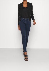 Blazer noir sur mesure avec fermetures éclair latérales sur un haut noir orné de dentelle, associé à un jean skinny bleu foncé et des sandales noires à talons.