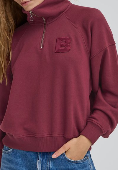 Burgundski pulover z visokim ovratnikom, pol-zapiranjem in vezenim 'B'. Ima ribane manšete in spodnji del. Material se zdi mehak in teksturiran.