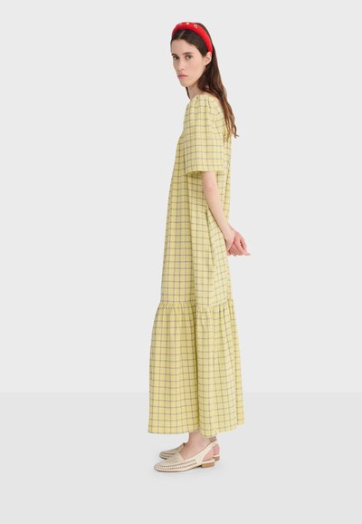 Femme de profil portant une robe midi à carreaux jaunes, des mocassins blancs en tissu tissé et un bandeau rouge rembourré avec des détails floraux.