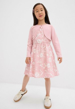 Ragazza giovane che indossa un vestito floreale rosa e un bolero coordinato, in piedi con una gamba in avanti e sneakers bianche con righe nere.