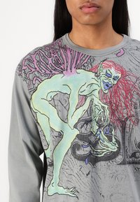 Šedá košile s dlouhým rukávem, zdobená surrealistickým grafickým potiskem v zelené, růžové a modré barvě, zobrazující humanoidní postavu s propracovanými detaily.