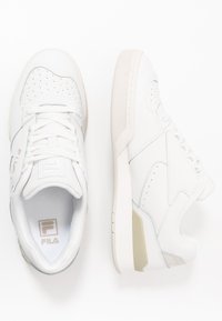 Sneakers bianchi realizzati in pelle liscia, con punte perforate, lacci piatti e un logo Fila discreto. Suole spesse di colore bianco sporco.