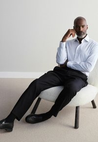 Chemise à rayures blanches et bleu clair, pantalon noir et mocassins. Assis sur une chaise crème à texture avec des pieds en bois foncé.