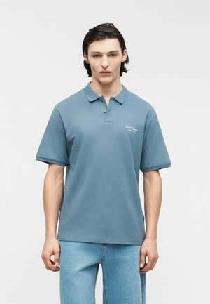 Mladý muž s tmavými vlasmi oblečený v tlmene modrej krátkorukávovej polo košeli a svetlomodrých džínsových nohaviciach, stojaci pred jednofarebným pozadím.