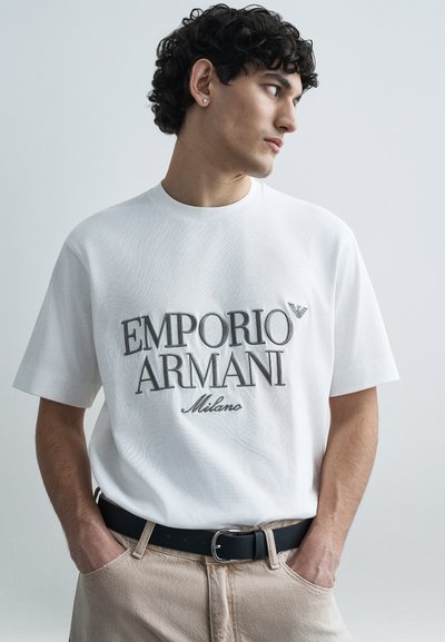 Λευκό βαμβακερό t-shirt με μεγάλο λογότυπο "EMPORIO ARMANI" σε γκρι, κοντά μανίκια και λαιμόκοψη στρογγυλή, σε συνδυασμό με μπεζ παντελόνι και μαύρη ζώνη.
