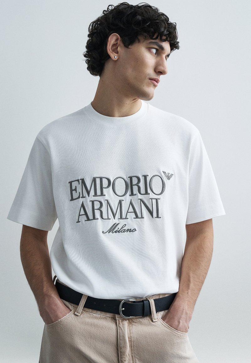 Biele bavlnené tričko s veľkým sivým logom "EMPORIO ARMANI", krátkymi rukávmi a okrúhlym výstrihom, v kombinácii s béžovými nohavicami a čiernym opaskom.