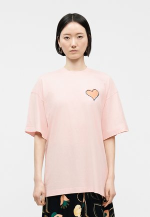 Vrouw met stijl zwart haar draagt een oversized lichtroze T-shirt met een hartapplicatie en een patroonrok tegen een effen lichte achtergrond.