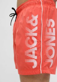 Crvene kupaće gaće s bijelim vertikalnim trakama i natpisom "JACK & JONES" sa strane, elastični pojas i crna vezica. Glatka tekstura tkanine.