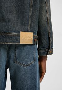 Mörk denimjacka med en beige läderlapp präglad med "HUGO." Har orangea sömmar och metallknappar som accent på ärmarna.