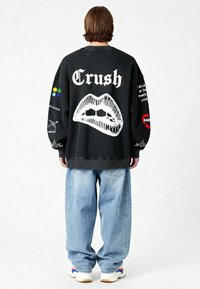 Felpa oversized nera con la scritta "Crush" in bianco sul retro, design di labbra grafiche e vari patch colorati sulle maniche. Indossata con jeans baggy blu chiaro.