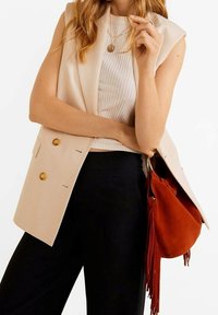 Beżowa kamizelka dwurzędowa z złotymi guzikami, noszona na białej bluzce z prążkami, w połączeniu z czarnymi spodniami i czerwoną zamszową torbą z frędzlami.