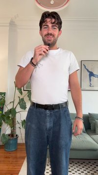 Bílé tričko s krátkým rukávem, volné modré džíny, černý pásek a náramky. Vnitřní prostředí s zelenou rostlinou a pohovkou v pozadí.