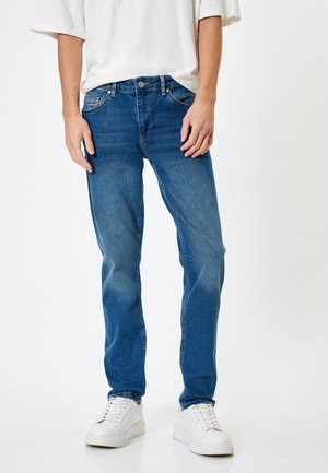 Vaqueros de denim azul con un corte recto, diseño de cinco bolsillos, ligero desgastado y cierre de botón plateado, combinados con zapatillas blancas.