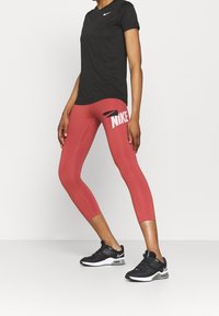 Camiseta negra de manga corta de Nike combinada con leggins capri rojos que presentan un gran logo de Nike en rosa, complementada con zapatillas deportivas negras.