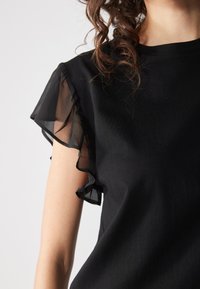 Haut noir avec des manches courtes en tulle volantes, présentant une texture de tissu lisse et une coupe décontractée. Pas de motifs visibles ni de détails en métal.