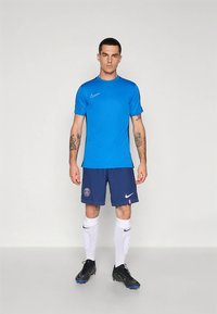 Mėlynas sportinis marškinėlis, derinamas su tamsiai mėlynomis šortais su logotipu, baltais kojinaitėmis ir juodais sportiniais bateliais. Audinys yra tekstūruotas ir drėgmę sugeriantis.