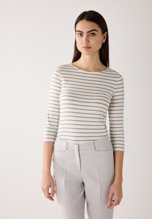 Femme aux longs cheveux foncés portant un haut blanc à rayures grises aux manches trois-quarts et un pantalon gris clair, se tenant devant un fond uni.