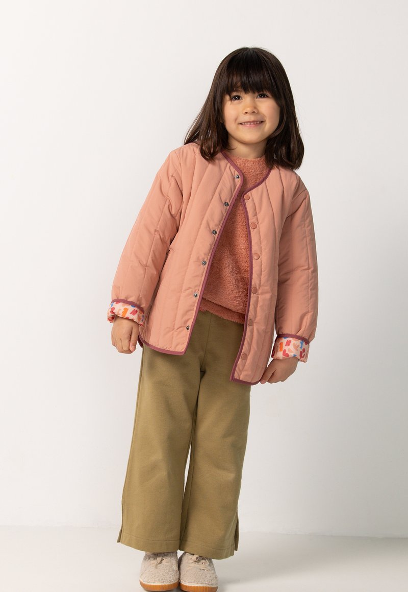 Jong meisje met donker haar, gekleed in een roze gewatteerde jas, roestkleurige trui, olijfgroene wijdlopende broek en beige schoenen, glimlachend en staand.