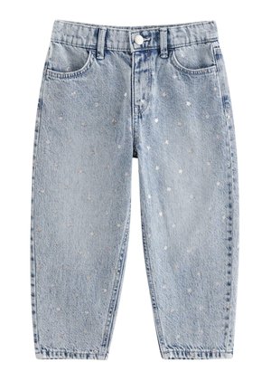 Barn med krøllet hår, smilende, iført en simpel hvid t-shirt, lyseblå jeans med små hvide mønstre og hvide sneakers, stående på et hvidt gulv.