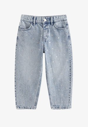 Barn med krøllet hår, smilende, iført en simpel hvid t-shirt, lyseblå jeans med små hvide mønstre og hvide sneakers, stående på et hvidt gulv.