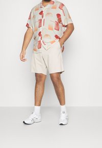 Camicia a maniche corte con bottone, caratterizzata da forme astratte rosse e arancioni su uno sfondo chiaro, abbinata a pantaloni beige e scarpe da ginnastica bianche.