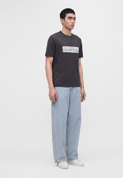Άνδρας στέκεται φορώντας μαύρο μπλουζάκι Emporio Armani, ανοιχτόχρωμο τζιν με φαρδιά μπατζάκια και λευκά αθλητικά παπούτσια, σε μονόχρωμο φόντο.