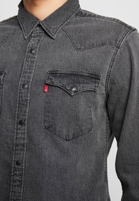 Lähikuva henkilöstä, joka käyttää haalistunutta mustaa farkkupaitaa, jossa on painepainikkeet ja läppätasku rinnassa, jossa on pieni punainen lipuke.