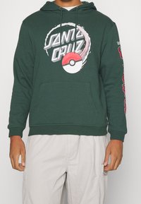 Hombre con sudadera oscura verde con el texto blanco "Santa Cruz" y un gráfico de Pokeball rojo-blanco, combinada con pantalones beige claros.