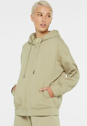 Woman with short blonde hair wearing a beige zip-up hoodie with hands in pockets, looking to the side against a plain background.