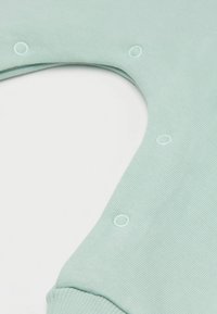 Jasnozielona, żebrowana tkanina z naszytymi zatrzaskami rozmieszczonymi wokół wygiętego otwarcia oraz z mankietem na krawędzi, na białym tle.