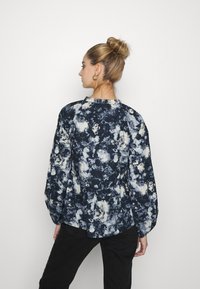 Blusa azul-marinho com padrões abstratos em cores claras, mangas bufantes longas e decote arredondado. O material parece leve com um brilho sutil.