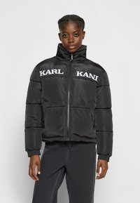 Giacca a vento nera con collo alto e zip frontale. Presenta i loghi "KARL" e "KANI" bianchi sul petto. Design imbottito con polsini a costine.