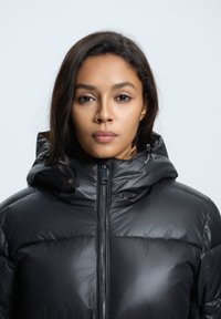Chaqueta puffer negra con cuello alto y capucha. Presenta un cierre frontal y una textura suave y brillante. Diseño minimalista sin patrones.