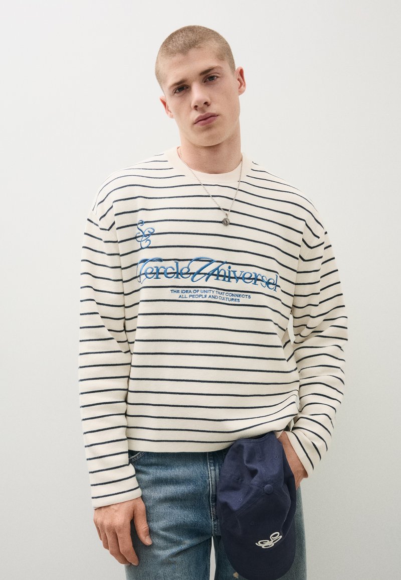 Jeune homme aux cheveux courts portant un pull rayé blanc et bleu marine avec du texte et tenant une casquette bleu marine dans une main.