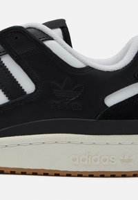 Zbliżenie na czarno-biały sneaker marki Adidas, widoczny bok z teksturowaną podeszwą, trzema paskami oraz wytłoczonym logo Adidas na skórzanym wierzchu.