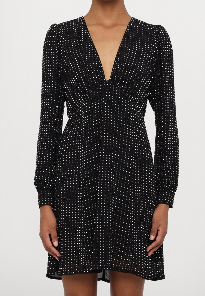 Robe noire à manches longues avec des pois blancs, décolleté plongeant en V et taille froncée, portée par une personne avec les mains détendues le long du corps.