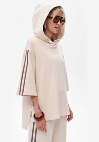 Beżowa bluza z kapturem o szerokim dekolcie, krótkich rękawach i czerwonych pionowych paskach po bokach. Posiada gładki materiał i luźny krój.