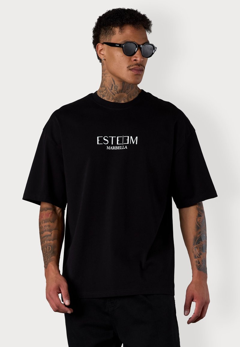 T-shirt noir oversize en coton, avec le texte blanc "ESTEEM MARBELLA" sur le devant. Porté avec des lunettes de soleil noires.