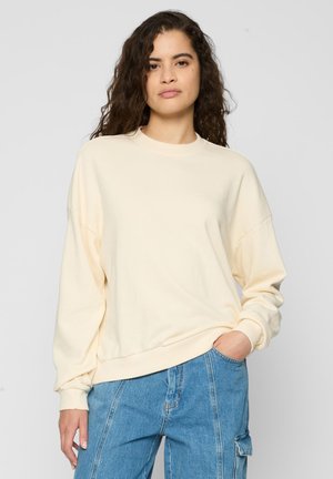 Sweatshirt crème avec un col rond, des manches longues, des épaules déchues et des poignets côtelés, associé à un jean baggy bleu clair.