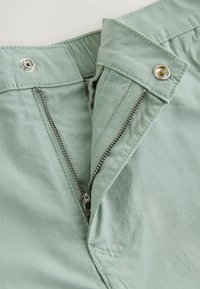 Pantalon vert clair avec une fermeture éclair en métal ouverte et une fermeture par bouton sur la ceinture, montrant des coutures détaillées et la texture du tissu.
