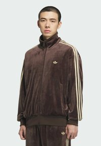 Young man wearing a brown velour Adidas tracksuit with beige stripes and logos on sleeves, chest, and pants, standing against a plain background.
