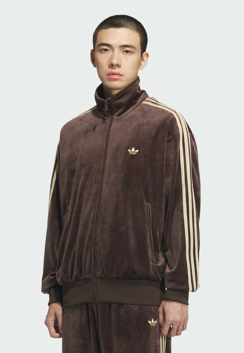Young man wearing a brown velour Adidas tracksuit with beige stripes and logos on sleeves, chest, and pants, standing against a plain background.
