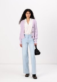 Lilla ribbestrikket cardigan over hvit bluse med blonder, kombinert med lyseblå dressbukser med vidde og svart håndveske. Brukt med svarte støvler.