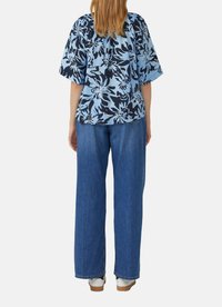 Person in einem blauen, blumigen Kurzarmoberteil und geraden blauen Jeans, steht mit dem Rücken zu einer weißen Hintergrund.