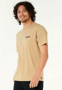 Beige T-shirt met korte mouwen van katoen met een ronde hals. Beschikt over kleine zwarte en rode "RIPCURL" tekst op de linkerkant van de borst.