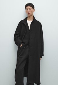 Cappotto lungo nero in materiale morbido con vestibilità rilassata, caratterizzato da bottoni, tasche laterali e colletto, indossato sopra un maglione scuro.