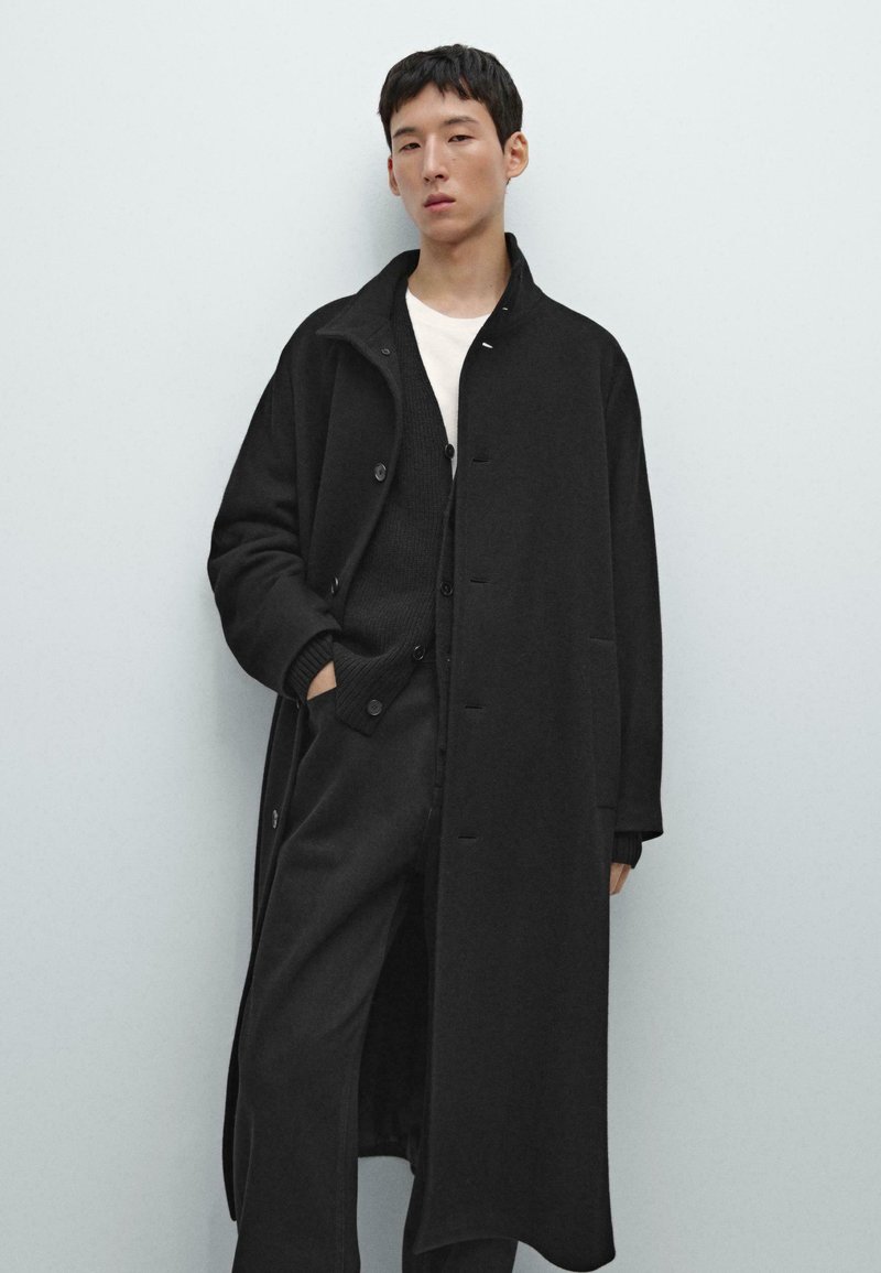 Cappotto lungo nero in materiale morbido con vestibilità rilassata, caratterizzato da bottoni, tasche laterali e colletto, indossato sopra un maglione scuro.